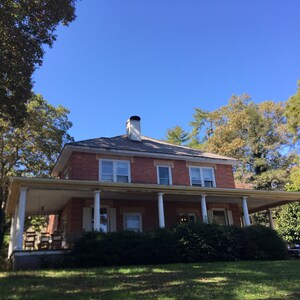 May include: A brick house with a large front porch and white columns. The house has a gray roof and is surrounded by trees.