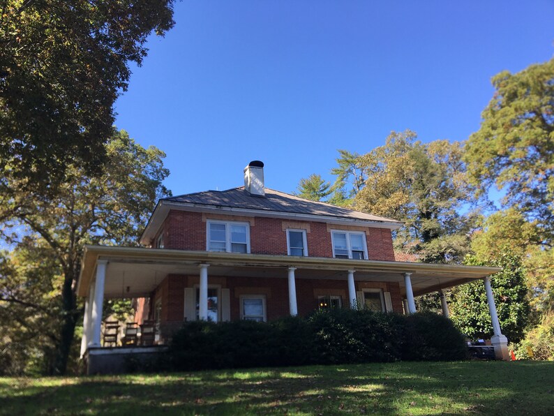 May include: A brick house with a large front porch and white columns. The house has a gray roof and is surrounded by trees.