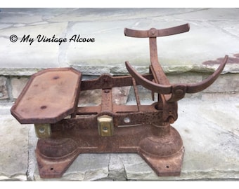 May include: A rusty antique metal scale with a weighing platform and a hanging beam. The scale is sitting on a stone surface.