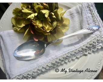May include: A silver spoon with an ornate design on a white linen napkin with a beaded trim. The spoon is resting on a white plate with a green flower.