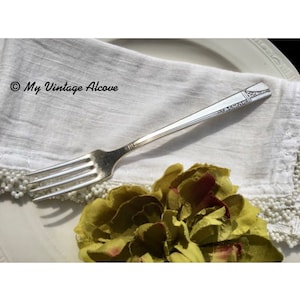 May include: A silver fork with an ornate design on the handle. The fork is resting on a white napkin with a beaded edge.