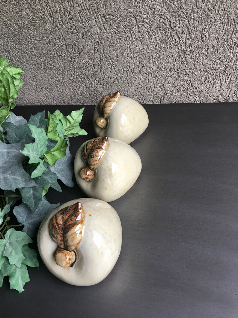 May include: Three ceramic apples with brown leaves, each with a glossy white finish. The apples are arranged in a row on a dark surface.