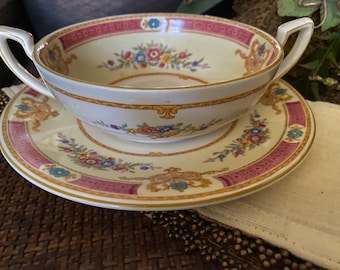 May include: A vintage cream-colored soup bowl with a floral pattern and gold trim. The bowl has two handles and is sitting on a matching saucer.