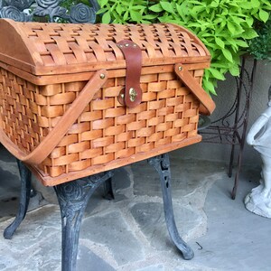 May include: A brown wicker picnic basket with a wooden lid and leather handles. The basket is sitting on a black metal stand with ornate legs.