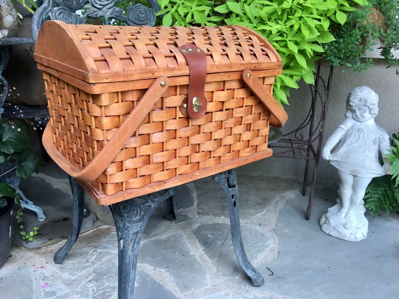 May include: A brown wicker picnic basket with a wooden lid and leather handles. The basket is sitting on a black metal stand with ornate legs.