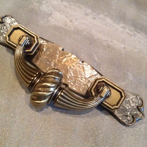 May include: A decorative cabinet pull with a brass finish. The pull features a rectangular backplate with ornate detailing and a curved handle. The handle has a ribbed design and is attached to the backplate with decorative hardware. The hardware is a mix of gold and silver tones.