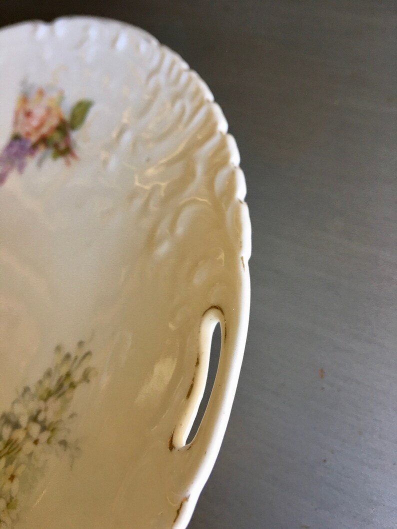 May include: White porcelain serving dish with a floral design and a gold-trimmed handle. The dish has a raised, textured rim.