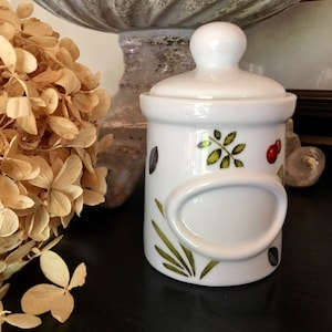 May include: White ceramic jar with a green leaf and red berry design. The jar has a blank oval label and a white lid.
