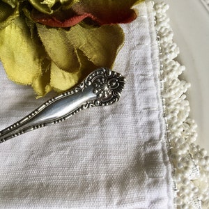 May include: A white linen napkin with a beaded edge and a silver spoon on top. The napkin is folded in half and the spoon is resting on the top fold. A green and red flower is in the background.