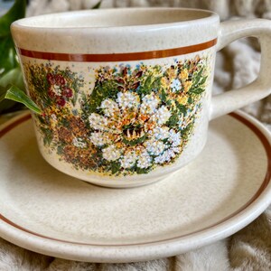 May include: A white ceramic teacup and saucer set with a floral pattern in shades of brown, green, and white. The cup has a brown rim and a handle. The saucer has a brown rim.
