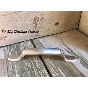 May include: A silver cabinet pull with a slightly curved design. The pull is on a weathered wooden surface.