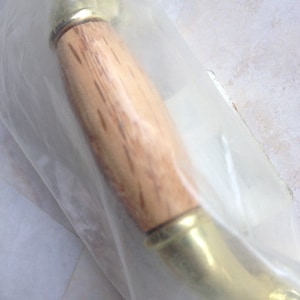 Puede incluir: Un tirador de puerta con un mango de madera clara y detalles metálicos de color latón. El tirador está parcialmente encerrado en una bolsa de plástico transparente.