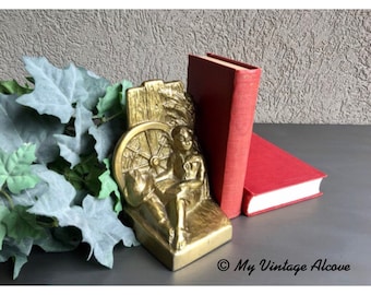 May include: A gold-colored metal bookend featuring a seated figure with a hat, a wagon wheel, and a red book. The bookend is placed next to a red book with white pages. The background is a textured gray wall.