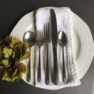 Puede incluir: Un juego de cubiertos de plata, que incluye un cuchillo, un tenedor y una cuchara, está dispuesto sobre una servilleta blanca con un borde de cuentas. Los cubiertos están colocados en un plato blanco con un borde decorativo.
