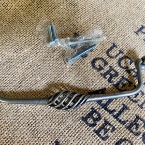 Könnte beinhalten: Ein silberner Metall-Schrankgriff mit einem gedrehten Design und abgerundeten Enden. Der Griff befindet sich auf einem Jute-Hintergrund.