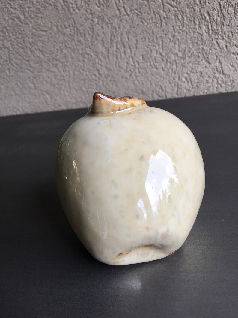 May include: A ceramic apple sculpture with a white glaze and brown accents. The apple is slightly misshapen and has a glossy finish.