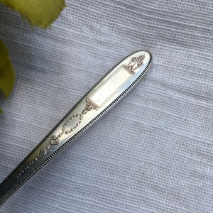 May include: Close-up of the top of a silver spoon with an ornate design. The spoon has a decorative handle with a raised oval and a beaded border.