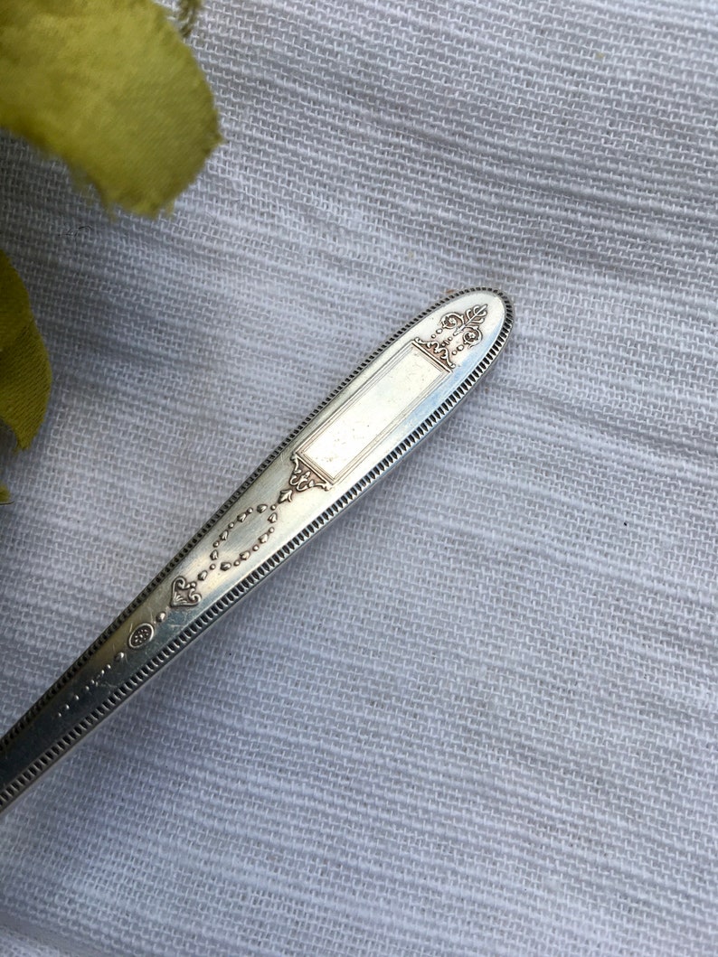 May include: Close-up of the top of a silver spoon with an ornate design. The spoon has a decorative handle with a raised oval and a beaded border.