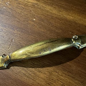 May include: A vintage brass drawer pull with a curved, elongated design. The hardware has a tarnished, aged appearance, with visible wear and tear. The pull features decorative end pieces and a central grip area, suggesting a classic or antique style.