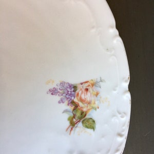 May include: White porcelain plate with a raised edge and a floral design. The design features a yellow rose with purple flowers and green leaves.