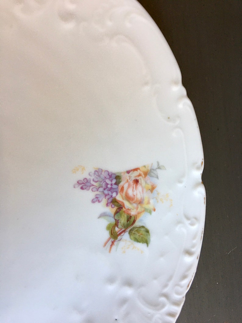 May include: White porcelain plate with a raised edge and a floral design. The design features a yellow rose with purple flowers and green leaves.