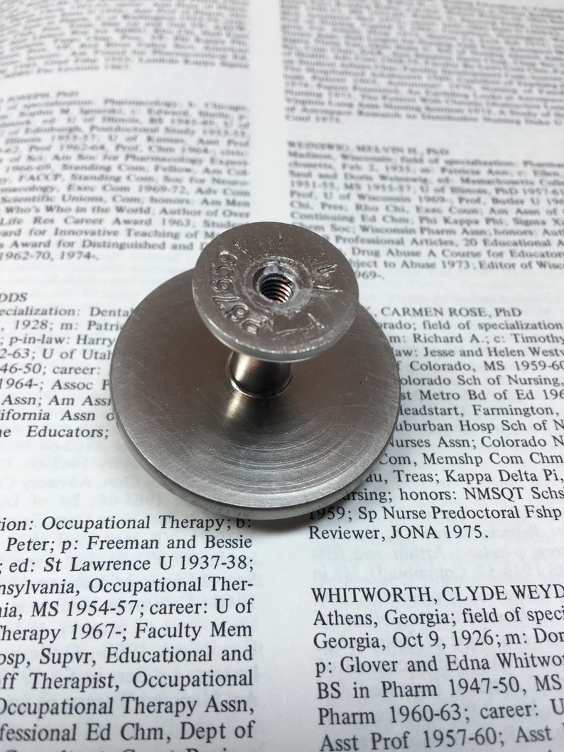 Op de afbeelding: Een zilveren metalen knop met een ronde basis en een schroef met schroefdraad. De knop heeft de gravure "Georgian" erop.