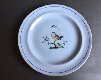 May include: A white ceramic plate with a blue rim features a hand-painted design of a yellow bird perched on a branch with green leaves and pink fruit.