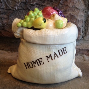 Peut inclure: Pot en céramique blanche en forme de sac avec un couvercle. Le couvercle est décoré d'un groupe de fruits et de raisins colorés. Le pot porte l'inscription "Home Made" sur le devant.