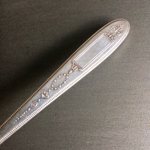 May include: Silverware handle with an ornate design and a blank rectangular space for engraving. The handle is silver in color and has a beaded edge.