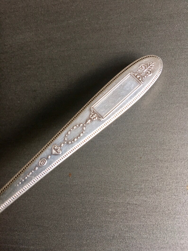 May include: Silverware handle with an ornate design and a blank rectangular space for engraving. The handle is silver in color and has a beaded edge.