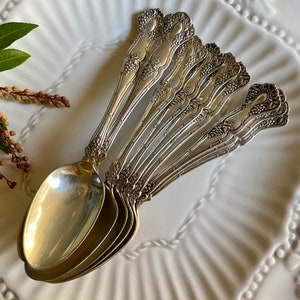 Puede incluir: Un juego de seis cucharas de plata con mangos adornados, que presentan un diseño floral. Las cucharas están dispuestas en forma de abanico sobre un plato blanco.