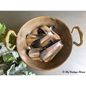 May include: A copper bowl with two ornate handles holds a pile of vintage metal shoe horns. The shoe horns are a mix of silver and copper tones.