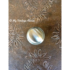 May include: A silver metal knob with a smooth, shiny surface. The knob is set against a brown background with a textured, embossed pattern.