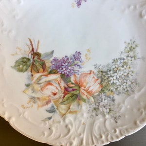 May include: A white porcelain serving platter with a raised rim and two handles. The platter is decorated with a floral design featuring yellow roses, purple and white lilac flowers, and green leaves.