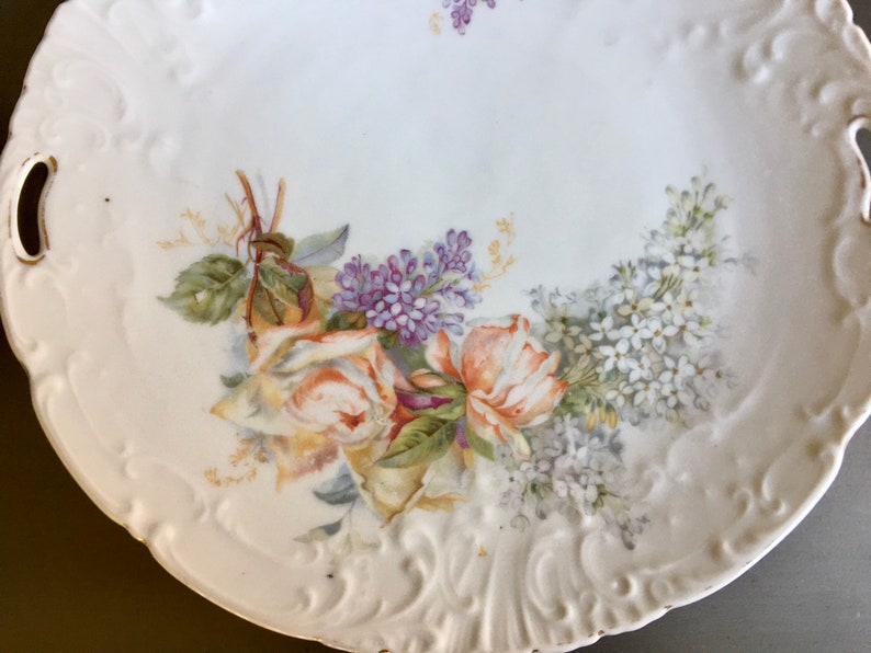 May include: A white porcelain serving platter with a raised rim and two handles. The platter is decorated with a floral design featuring yellow roses, purple and white lilac flowers, and green leaves.