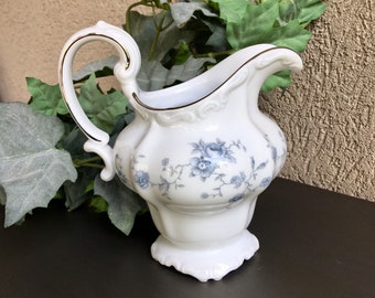 May include: A white porcelain creamer with a blue floral pattern and gold trim. The creamer has a delicate handle and a scalloped base.