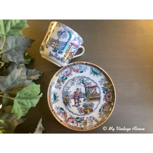 May include: A white teacup and saucer with a blue and gold floral pattern. The pattern features a pagoda and figures in a garden setting.