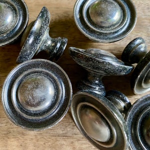 May include: A group of eight black and silver cabinet knobs with a circular design. The knobs have a raised center and a textured finish.