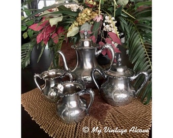 May include: A silver tea set with a teapot, creamer, and sugar bowl. The set has an ornate design and is sitting on a brown fringed table runner.
