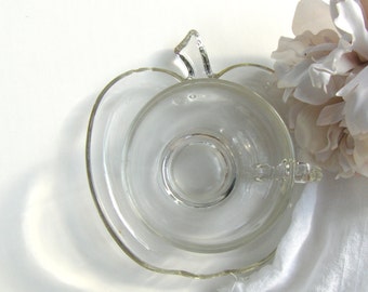 May include: A clear glass apple-shaped teacup and saucer set. The cup has a handle shaped like a leaf and the saucer has a scalloped edge.