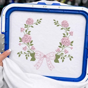May include: A white fabric piece secured in a blue rectangular embroidery hoop, featuring a machine-embroidered design of two symmetrical floral branches with light pink hydrangea blossoms, green leaves, and a central pink decorative bow.