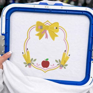 Peut inclure: Un morceau de tissu blanc fixé dans un tambour à broder bleu présentant un motif brodé composé d'un nœud jaune, de crayons jaunes et d'une pomme rouge centrés dans un cadre festonné décoratif jaune et rose sur le thème de l'école.