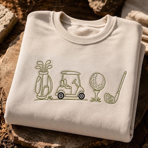 Könnte beinhalten: Ein cremefarbenes Sweatshirt mit Rundhalsausschnitt und olivgrüner Stickerei auf der Vorderseite. Das Design zeigt eine Golftasche, ein Golfcart, einen Golfball auf einem Tee und einen Golfschläger, die horizontal in der Mitte angeordnet sind.