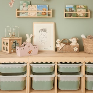 May include: A nursery scene with wooden shelves holding books and toys. A framed alphabet print and a drawing are on the wall. A pink toy basket and a wooden toy are on a wooden table. The color scheme is soft green and natural wood.