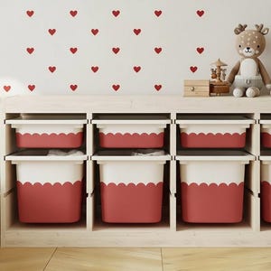 May include: A light wood storage unit with eight red and white bins. The bins have a scalloped design. A deer plush toy, a small wooden box, and a stack of books sit on top. Red heart decals decorate the white wall.