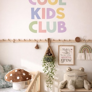 May include: A children's room with a wooden crib, a mushroom-shaped pillow, and a large stuffed frog. The wall features the words "COOL KIDS CLUB" in pastel colors, along with decorative items like a macrame rainbow and a framed quote.