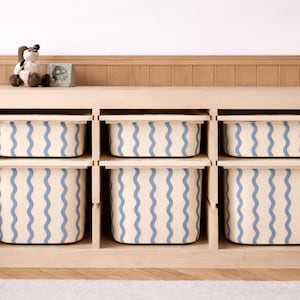 May include: A light wood storage unit with six cubbies, each holding a cream-colored bin with blue wavy lines. A stuffed dog and small framed picture sit on top. The unit is on a light wood floor.