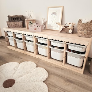 Può includere: Un mobile contenitore in legno per giocattoli con contenitori bianchi e un bordo smerlato. L'unità è sormontata da cesti, una stampa incorniciata e peluche. Un tappeto a forma di margherita è sul pavimento.
