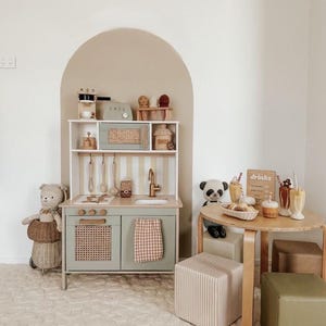 Può includere: Una cucina giocattolo per bambini in verde salvia con paraschizzi a righe e anta in tessuto. Il set include un tostapane giocattolo, una macchina per il caffè e vari utensili da cucina. Un tavolino con bevande e un panda sono presenti nella scena.