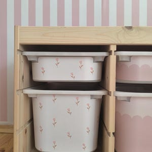 Peut inclure: Unité de rangement en bois avec deux bacs en plastique blancs décorés de motifs floraux roses. Les bacs sont empilés à l'intérieur de l'unité. L'arrière-plan présente un mur à rayures roses et blanches.